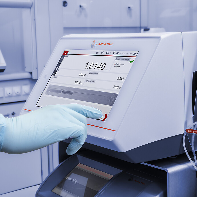 A gloved hand operates the touchscreen of an Anton Paar DMA 4500 M density meter with Xsample 530 sample changer in a lab setup.