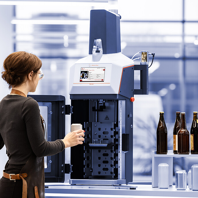 Technician loading a beverage can into the Anton Paar TPO 5001 Total Package Oxygen Meter; various bottles and cans shown for compatibility.